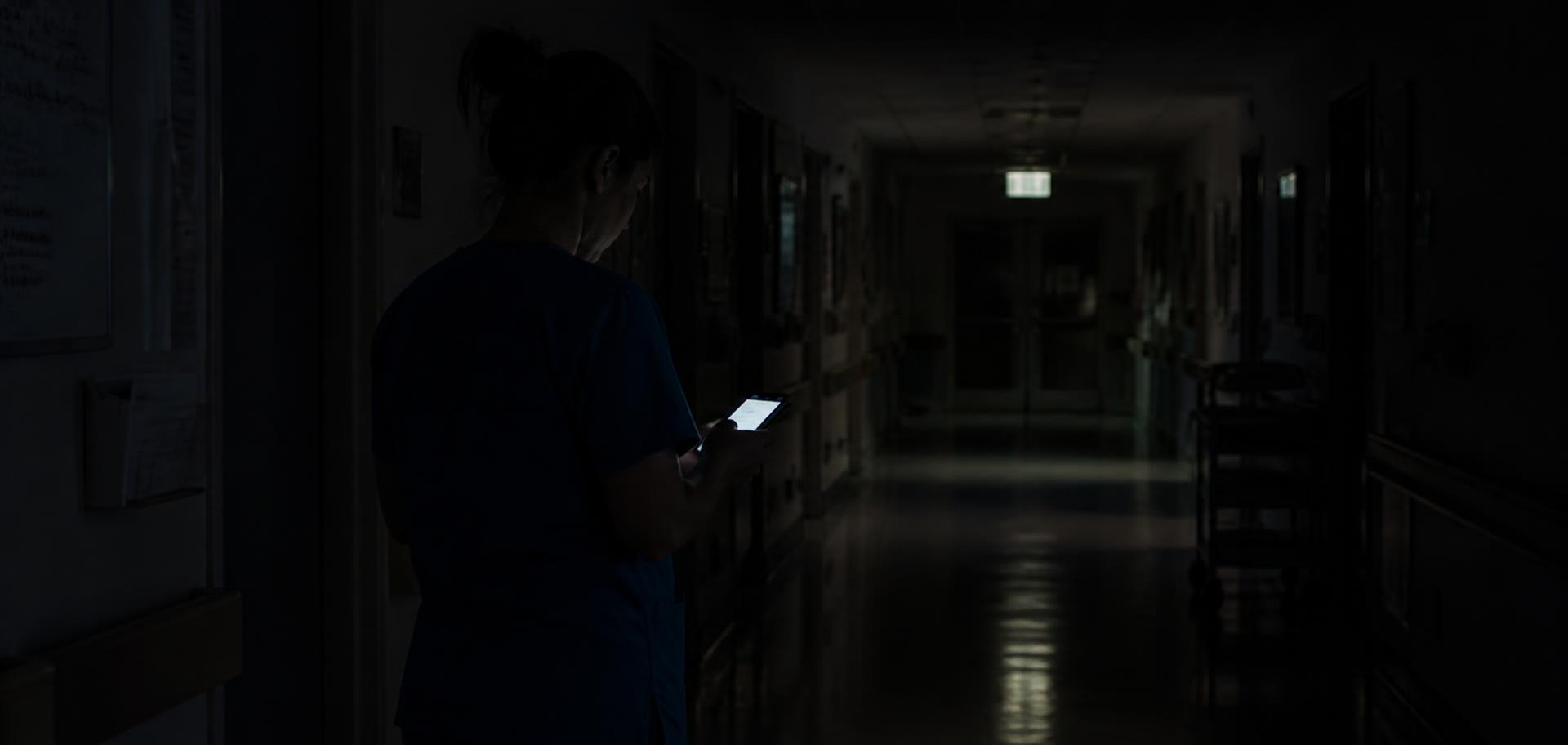 A nurse in scrubs reading a phone in a dim hospital corridor at night